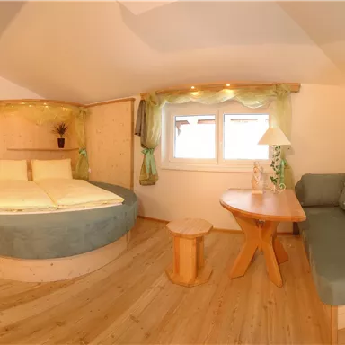 Ein gemütliches Zimmer mit einem runden Bett und großen Fenstern. Die Einrichtung ist hell und modern, mit einem kleinen Tisch und einer gemütlichen Sitzecke.