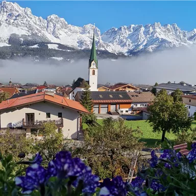 A picturesque mountain landscape with snow-covered peaks in the background. In the foreground, there is a small village with a church and colorful flowers.