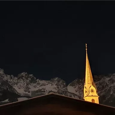 An impressive mountain landscape at night, with a illuminated church steeple in the foreground. The sky is dark and the mountains are partially snow-covered.