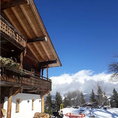 A cozy wooden house in the snow, surrounded by snow-covered trees. The sky is bright blue with some clouds in the background.