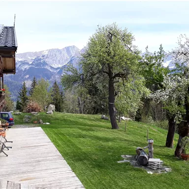 Eine malerische Gartenlandschaft mit blühenden Bäumen und weitem Blick auf die Berge. Im Vordergrund befindet sich eine Holzterrasse.