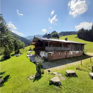Ein gemütliches Holzhaus umgeben von grünem Rasen und Bäumen. Im Hintergrund sind sanfte Hügel und ein blauer Himmel mit wenigen Wolken zu sehen.