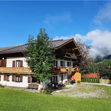 A traditional alpine house with a balcony and colorful flowers. The surroundings are green with mountains and clouds in the background.