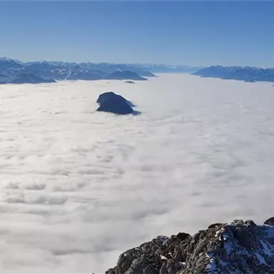 Ein atemberaubender Blick auf schneebedeckte Berge, die aus einer dichten Wolkendecke ragen. Der Himmel ist klar und blau, was die Landschaft noch majestätischer erscheinen lässt.