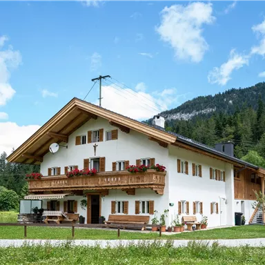 A charming, traditional house in the mountains, surrounded by green meadows. The sky is clear with a few clouds, and the view of the surrounding mountains is beautiful.