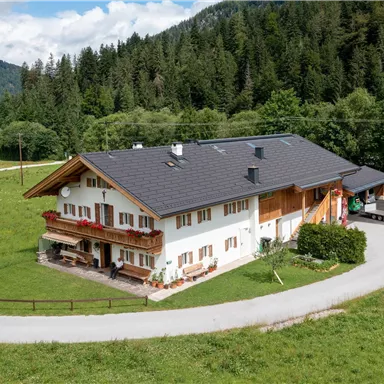 A large, modern house surrounded by green grass and trees. The landscape is characterized by hills and a clear sky.