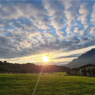 A picturesque sunset over a green field, surrounded by mountains. The sky is filled with scattered clouds in soft colors.