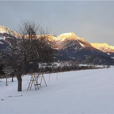 A winter landscape with snowy fields and majestic mountains in the background. A tree and a swing are visible in the foreground.
