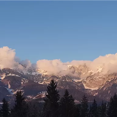 A majestic mountain range with snow-covered peaks and clouds. The clear sky and the surrounding trees create a tranquil atmosphere.