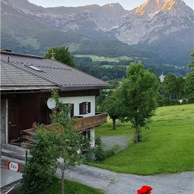 A picturesque house in the mountains with a beautiful view of the surrounding peaks. The garden is lush green and adorned with red flowers.
