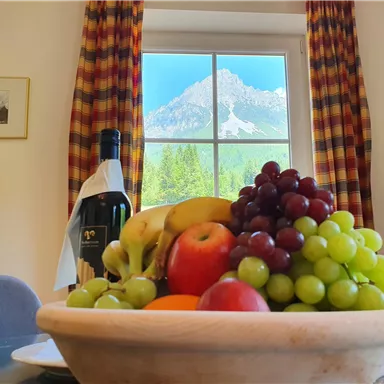 A bowl of fresh fruit, including apples, grapes, and bananas, sits on a table. In the background, there is a window with a stunning mountain landscape.