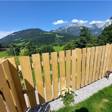 A wooden plank fence overlooking green meadows and mountains. The sky is clear and blue, creating a beautiful landscape.