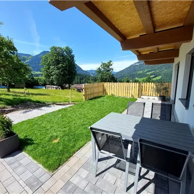 A beautiful outdoor area with a terrace, a table, and chairs. In the background, green meadows and mountains can be seen.