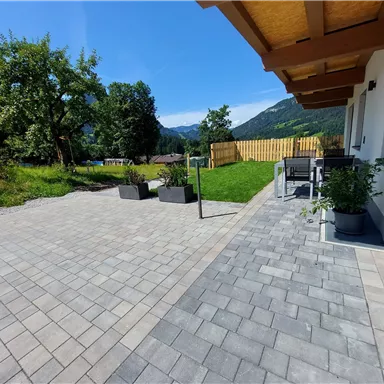 A beautiful outdoor area with paved flooring and modern furniture. In the background, trees and mountains are visible under a clear blue sky.