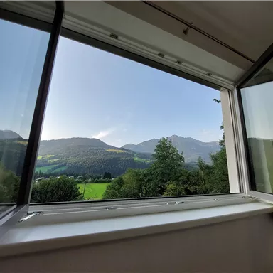 An open window overlooking green meadows and mountains. The sky is clear and the landscape is calm and idyllic.