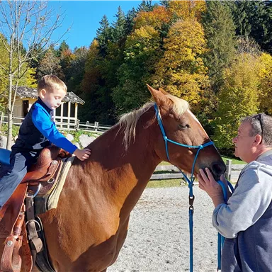 A boy is sitting on a horse and talking to an adult. In the background, there are trees with colorful foliage and a riding station visible.