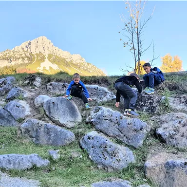 Three children are playing and climbing on a rocky hillside. In the background, majestic mountains and a clear sky can be seen.