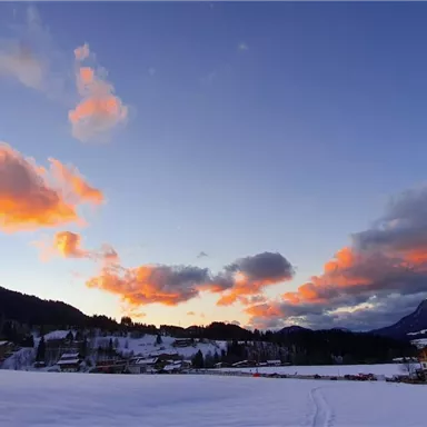 A snowy landscape with mountains and houses in the foreground. The sky is streaked with orange clouds.