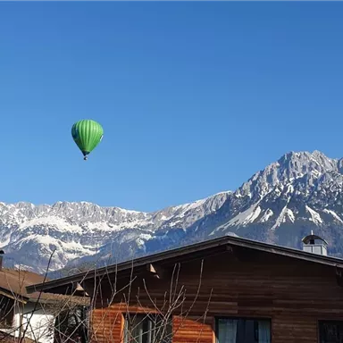A hot air balloon floats over snow-covered mountains, surrounded by a clear blue sky. In the foreground, traditional wooden buildings can be seen.