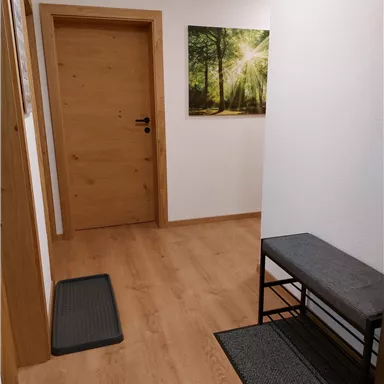 Ein heller Flur mit Holzfußboden und einer braunen Tür. An der Wand hängt ein Bild von einem sonnigen Wald.