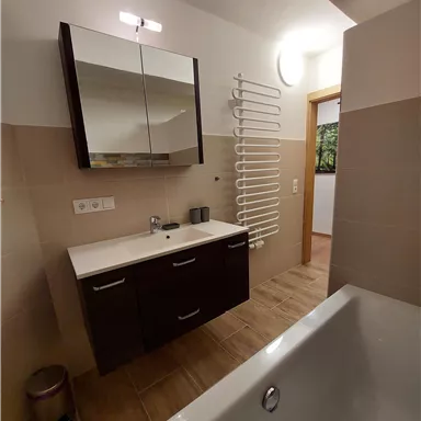 Ein modernes Badezimmer mit einem Waschbecken, einem großen Spiegel und einer Badewanne. Die Wände sind hell gefliest und es gibt einen Handtuchwärmer.