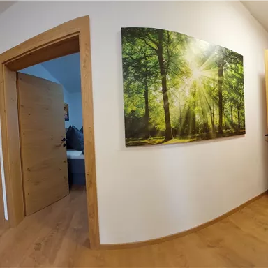 Ein heller Flur mit Holzfußboden und offenen Türen zu mehreren Räumen. An der Wand hängt ein Bild von einem sonnendurchfluteten Wald.