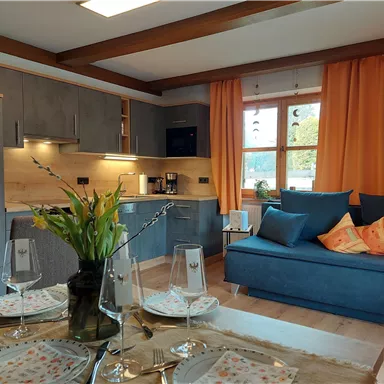 A modern kitchen with gray cabinets and a cozy living area. Orange curtains hang before a large window, and the table is lovingly set.