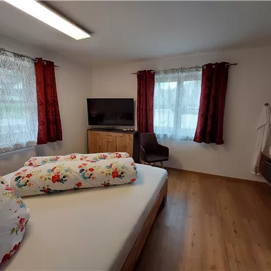 A cozy bedroom with a large window and red curtain. There is a bed with floral bedding and a TV on a wooden cabinet.