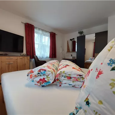 A cozy bedroom with a large bed and colorful pillows. In the background, there is a television and windows with red curtains.