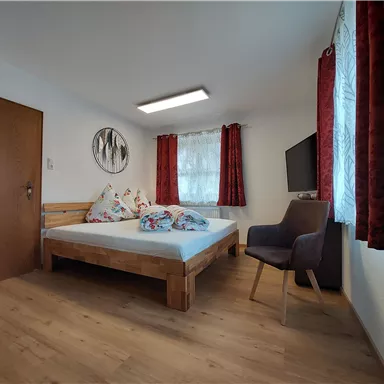 A cozy bedroom with a wooden bed and colorful pillows. The room has red curtains and a TV on the wall.