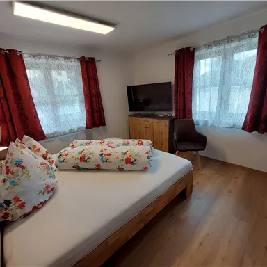 A cozy bedroom with a wooden bed and colorful pillows. Red curtains hang in front of the windows, and a TV is on a wooden dresser.