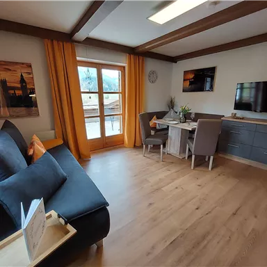 A cozy living room with a sofa, a dining table, and a television. Large windows let in a lot of light and offer a view outside.