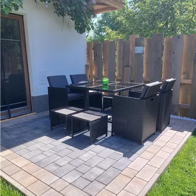 Ein gemütlicher Außenbereich mit einem Tisch und Stühlen aus Rattan. Der Bereich ist von einer Holzwand umgeben und hat grünes Gras.