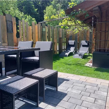 Ein gemütlicher Außenbereich mit einem Tisch und Stühlen aus schwarzem Geflecht. Im Hintergrund sind Liegestühle und ein kleiner Holzschuppen sichtbar, umgeben von grünem Rasen.