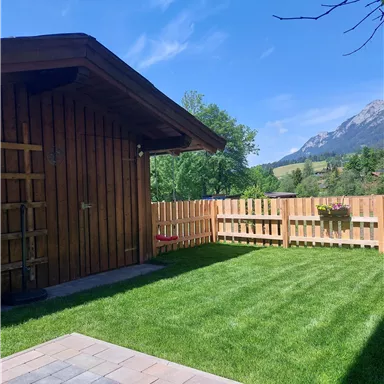 Ein gemütliches Holzhaus mit einem gepflegten Garten. Im Hintergrund sind grüne Bäume und Berge unter einem blauen Himmel zu sehen.