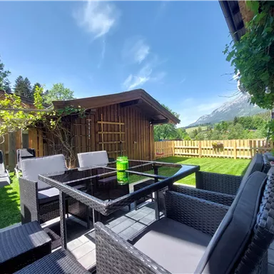 Ein gemütlicher Außenbereich mit modernen Möbeln und einem klaren blauen Himmel. Im Hintergrund sind Grünflächen und die Berge zu sehen.