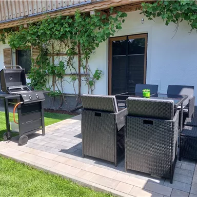 Eine gemütliche Terrasse mit einem Grill und modernen Gartensesseln. Im Hintergrund sind grüne Pflanzen und eine helle Hauswand zu sehen.