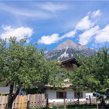 Ein malerisches Haus umgeben von grünen Bäumen und einer beeindruckenden Berglandschaft. Der Himmel ist klar mit einigen weißen Wolken.