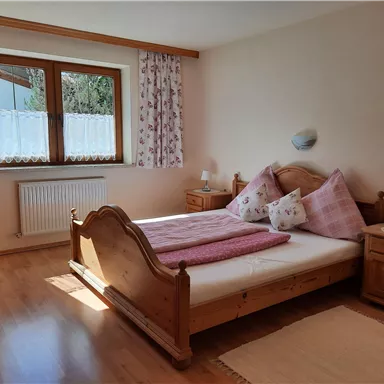 A cozy bedroom with a large bed and wooden dressers. The windows let in plenty of daylight, creating a bright atmosphere.