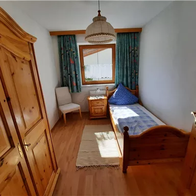 A cozy bedroom with wooden closets and a single bed. The room has a large window with curtains and a small bedside table.