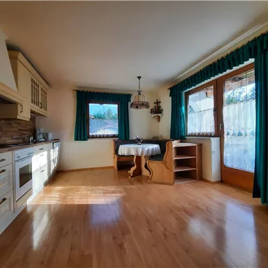 A bright and cozy kitchen with wooden floor. There is a table, chairs, and large windows with green curtains.