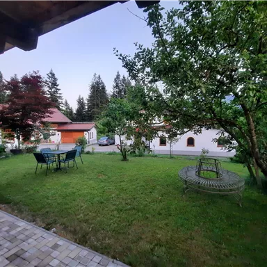 A beautiful garden with plenty of greenery and a table with chairs. In the background, there are some houses and trees visible.