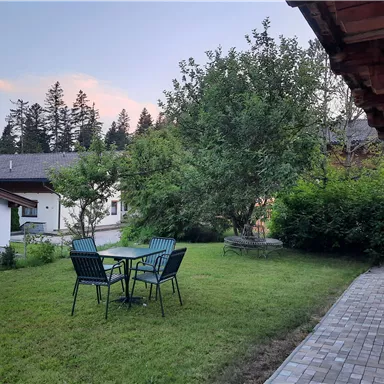 A quiet garden with a table and chairs, surrounded by trees and bushes. In the background, a few buildings can be seen.