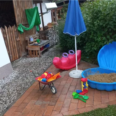 An outdoor play area with a sandbox, shovels, and a colorful slide. Backgrounds are green and a sun umbrella is ready.