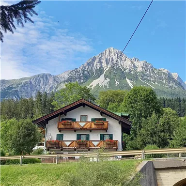 A charming house in the Alps with balconies and a green environment. In the background rises a majestic mountain under a blue sky.