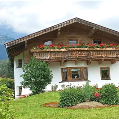 A beautiful, traditional house with a wooden balcony and colorful flowers in the garden. In the background, mountains and a green landscape can be seen.