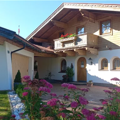A beautiful, traditional house with wooden cladding and a balcony. In front of the house, colorful flowers bloom in a well-maintained garden.