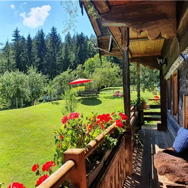 A cozy balcony with colorful flowers and a view of a green meadow. In the background, there are trees and a sun umbrella.