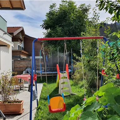 A beautiful garden with a swing and a slide. In the background, a trampoline and some green plants can be seen.