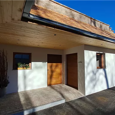 Das Bild zeigt den Eingang eines modernen Hauses mit einer hellen Fassade und einem Schindeldach. Der Himmel ist klar und blau, was eine angenehme Atmosphäre schafft.